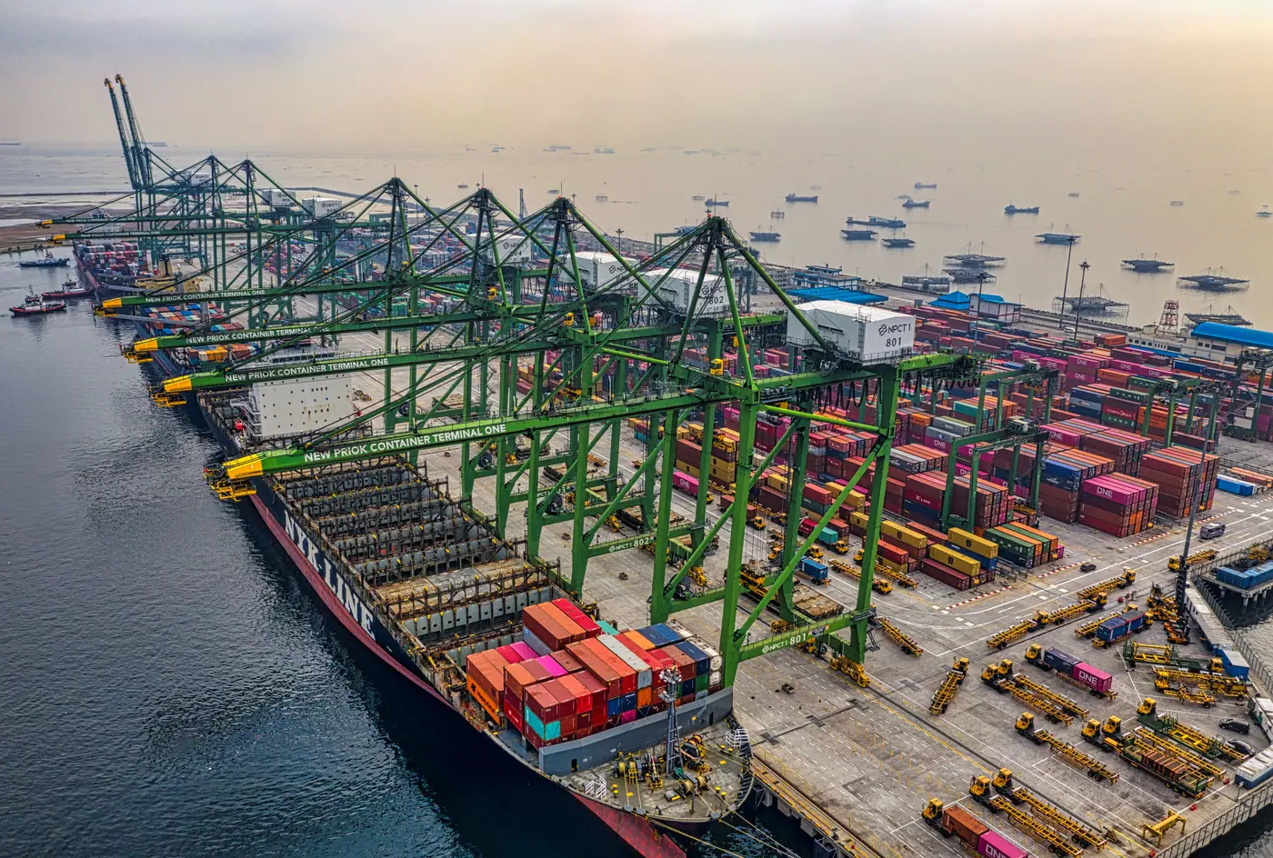 Busy cargo port in North Jakarta with cranes and container ships under a hazy sky.